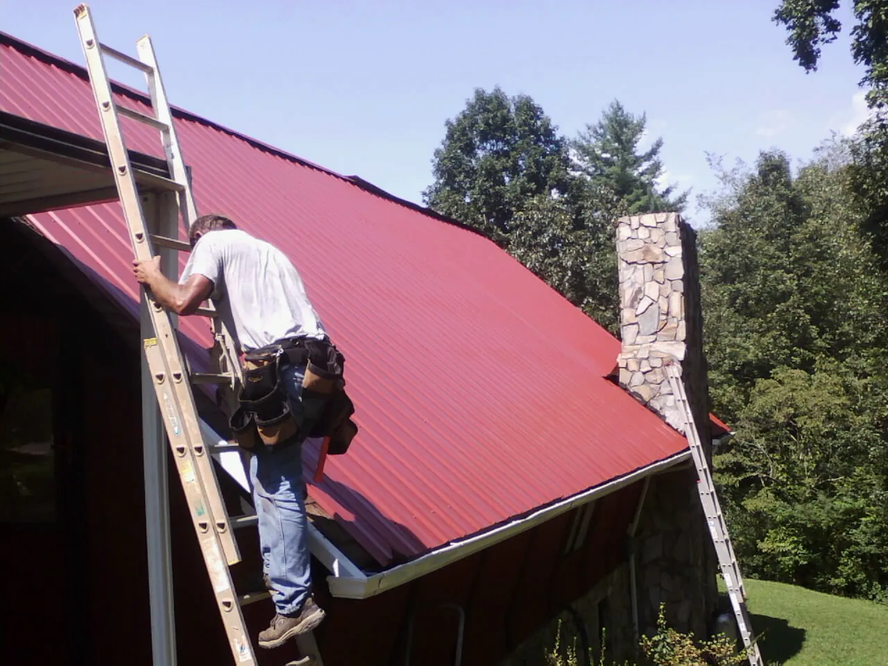 Roofer on a ladder beside a red metal roof and stone chimney — Soffit & Fascia Repair by Roofing Brothers in Allardt